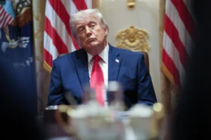 Person in a blue suit and red tie seated at a table, with American flags and ornate gold decoration in the background.