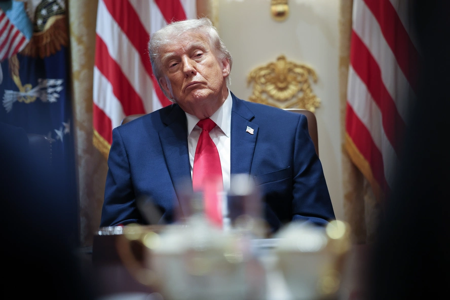 Miami Federal Judge Questions Legality of Trump’s $1.8 Billion IRS Settlement Person in a blue suit and red tie seated at a table, with American flags and ornate gold decoration in the background.