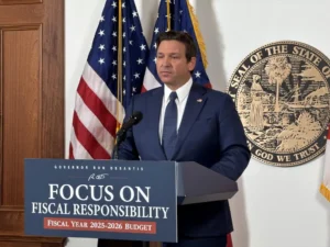 Person in suit standing at podium with "Focus on Fiscal Responsibility" sign, U.S. flags, and state seal in background.