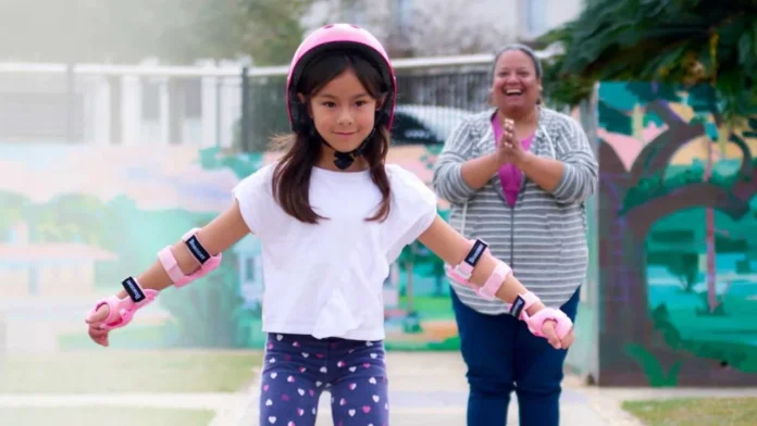 A child wearing protective gear roller-skates while an adult watches and applauds.